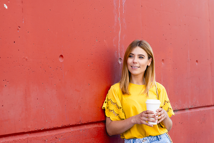 A young woman with her morning coffee, leaning against a red wall, by Cavan Images / rafa fernandez
