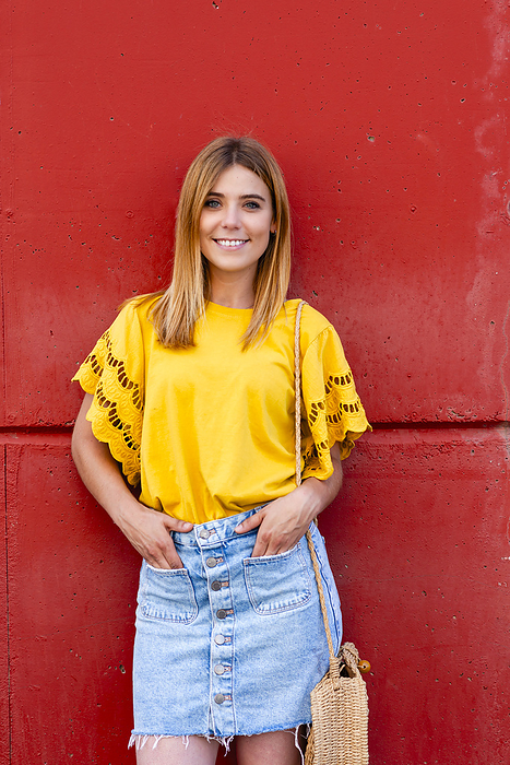 A young woman stands confidently against a red wall, her yellow blouse, by Cavan Images / rafa fernandez