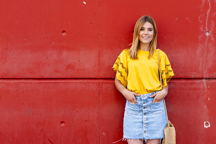 A young woman stands confidently against a red wall, her yellow blouse, by Cavan Images / rafa fernandez