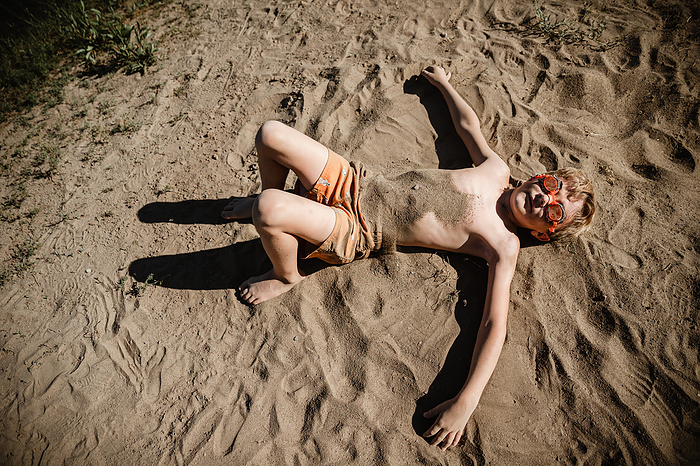 Young boy laying on beach covered in sand on summer day, by Cavan Images / Krista Taylor