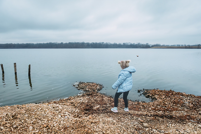 Child throwing rocks into water on shore of lake, by Cavan Images / Krista Taylor