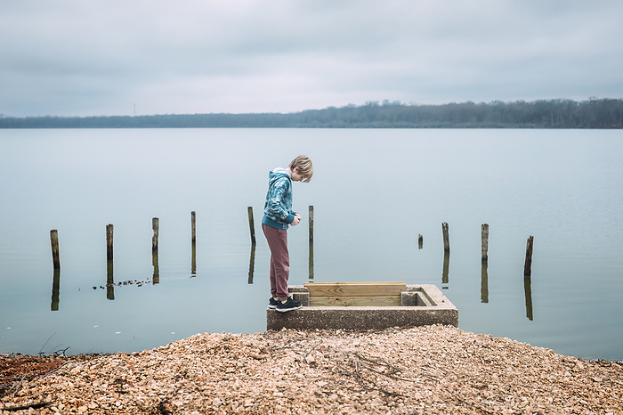 Boy looking into spillway on shore of lake on cloudy winter day, by Cavan Images / Krista Taylor