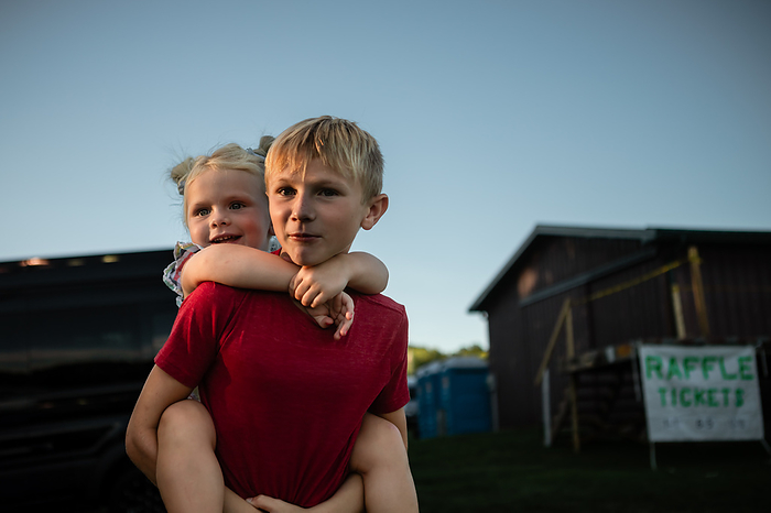 Brother giving little sister piggyback ride at summer festival, by Cavan Images / Krista Taylor
