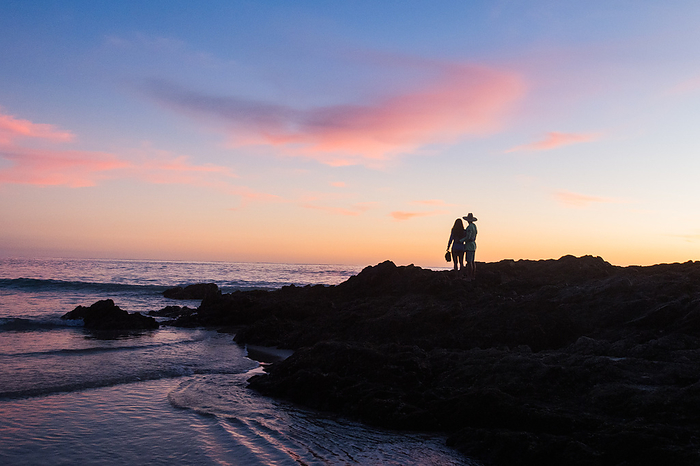 Two people stand on rocks at the beach at sunset., by Cavan Images / Michael Hanson