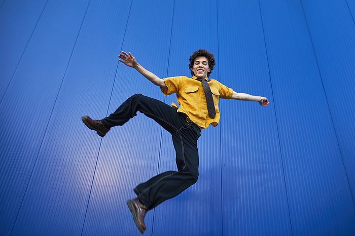 cheerful young man in a yellow shirt jumping on a blue background, by Cavan Images / Elena Perevalova