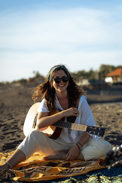 Woman musician sings and plays guitar on the beach. In a bandana. Bali, by Cavan Images / Yuliya Kirayonak