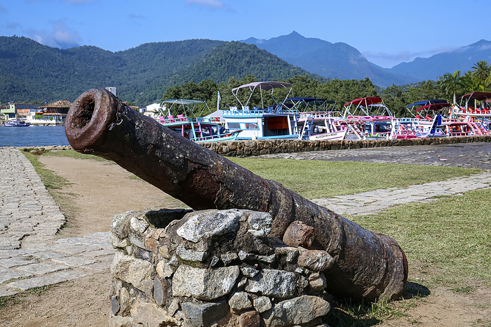 Close up of an old rusty cannon in front of an open space with grass, colorful boats and rainforest Close up of an old rusty cannon in front of an open space with grass, colorful boats and rainforest, by Zoonar Uwe Bergwitz