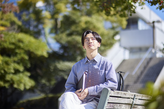 A male Japanese university student sitting on a bench on a university campus (People)