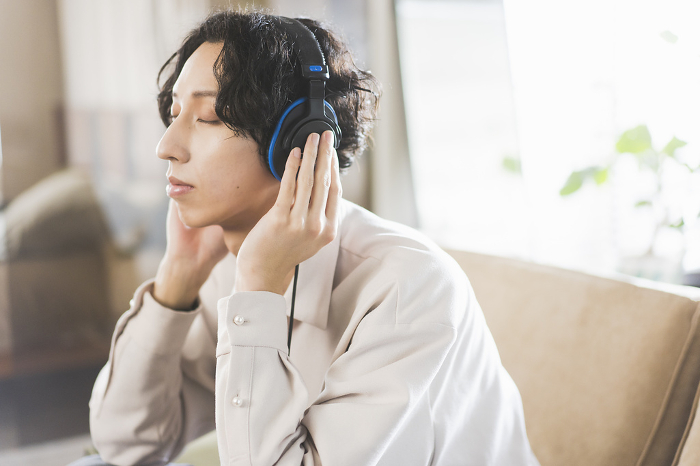Young Japanese man sitting on sofa at home listening to music with headphones / Generation Z (People)