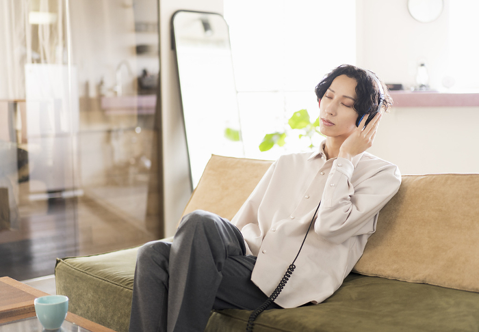 Young Japanese man sitting on sofa at home listening to music with headphones / Generation Z (People)