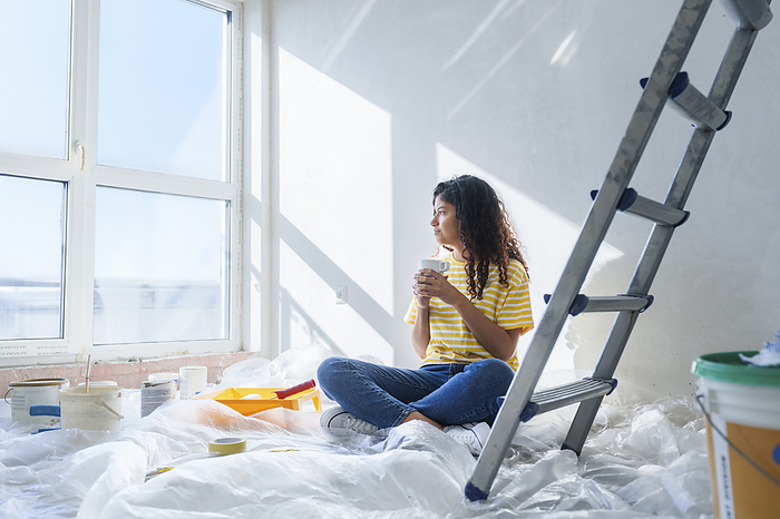Young woman with coffee cup sitting near ladder at new home