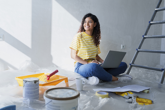 Smiling woman sitting on floor with laptop in room under renovation