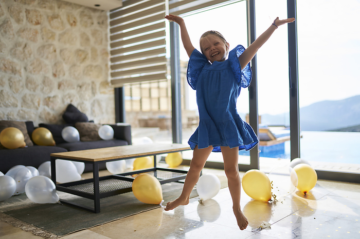 Happy girl jumping with arms raised by balloons on floor at villa