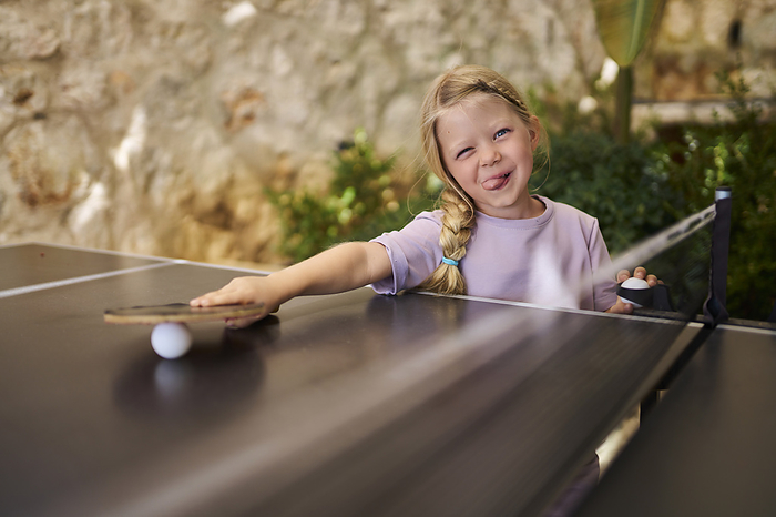 Playful girl sticking out tongue holding table tennis racket at villa