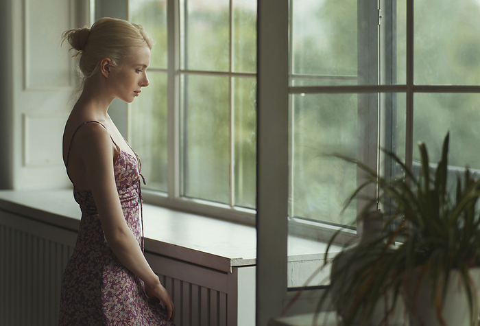 Young pensive blond woman standing next to the window Thoughtful woman standing near window