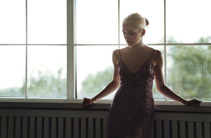 Young pensive blond woman standing next to the window Thoughtful young woman standing near window at home