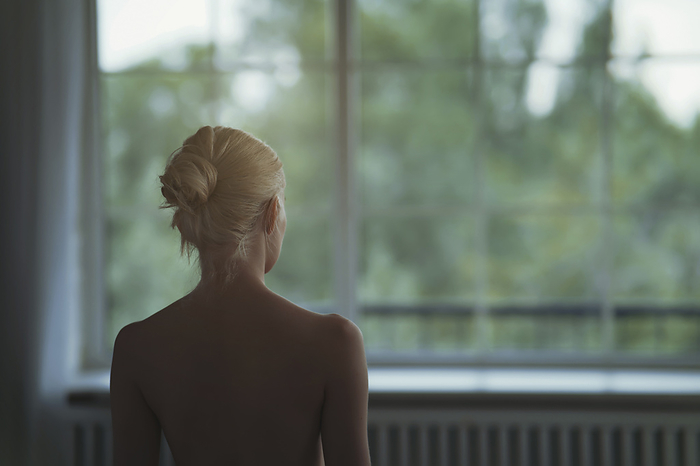 Young blond woman looking to the window Naked young woman with blond hair looking through window