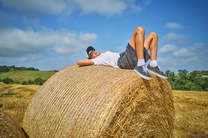 Boy lying on hay bale in field under cloudy sky