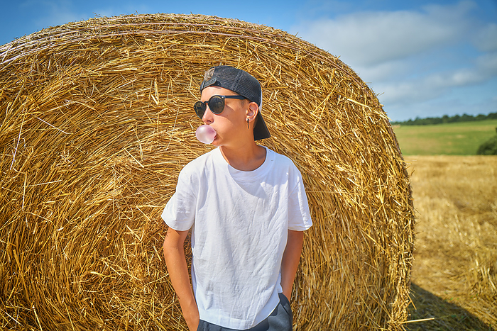Boy blowing bubble gum and leaning on bale of straw in field
