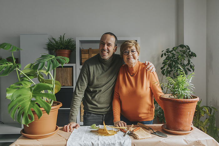 Smiling son with arm around mother standing near plants on table at home