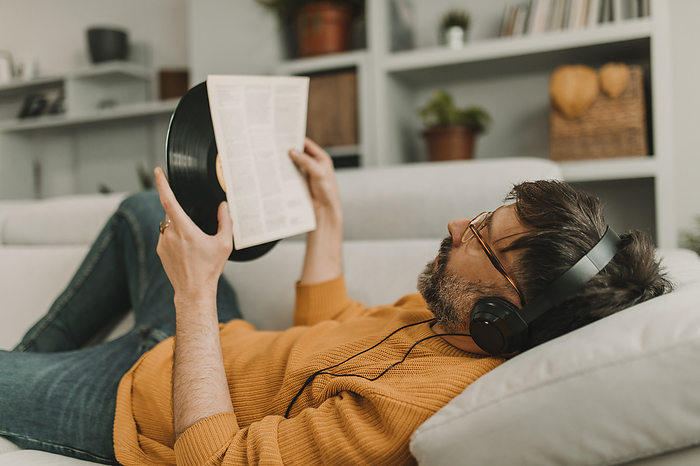 Man listening to music through headphones reading paper holding vinyl record at home