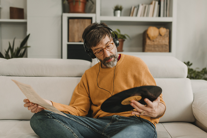 Man listening to music and reading paper holding record sitting on sofa at home