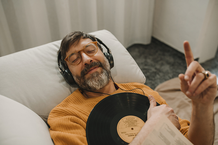 Man listening to music through headphones lying with record on sofa at home
