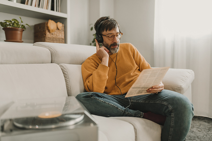 Man listening to music through headphones reading paper sitting by turntable on sofa at home