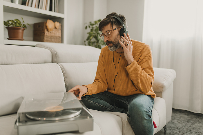 Man listening to music through headphones sitting by turntable on sofa at home