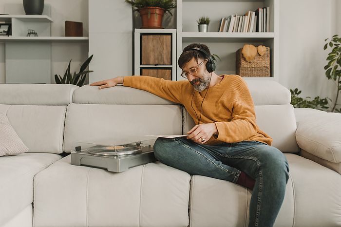 Man listening to music through headphones sitting by turntable on sofa in living room