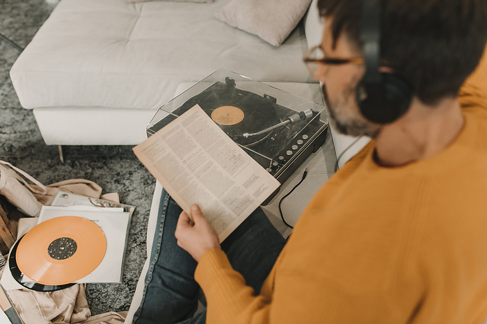 Man listening to music through headphones reading paper sitting by record player on sofa at home