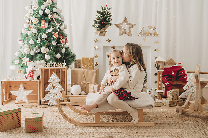 Girl kissing sister on sled near Christmas decoration at home
