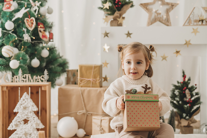 Cute girl sitting with Christmas present near decoration at home