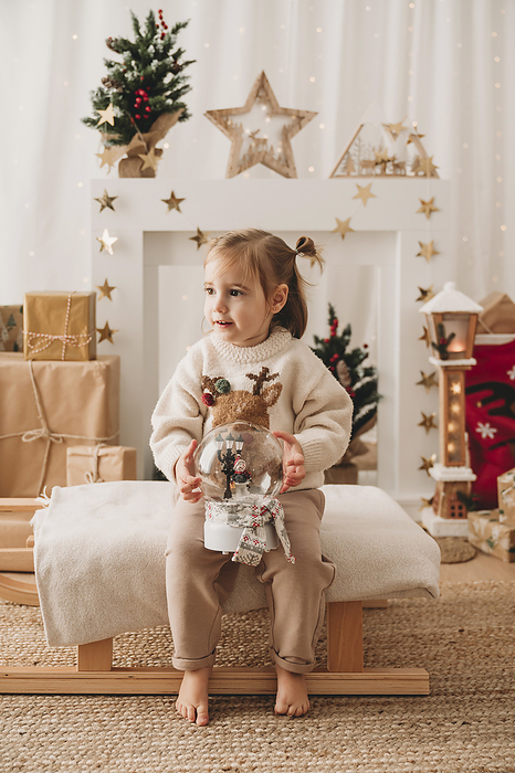 Girl with snow globe sitting near Christmas decoration at home