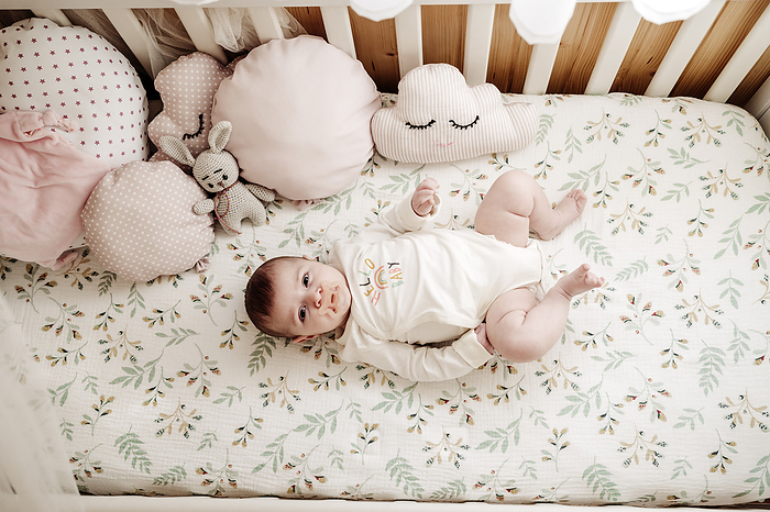Baby girl lying in crib near cushions at home