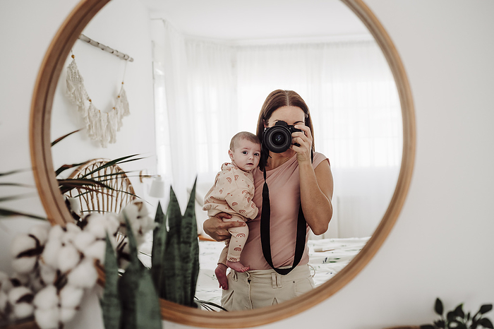 Mother carrying baby girl and taking picture through camera at home