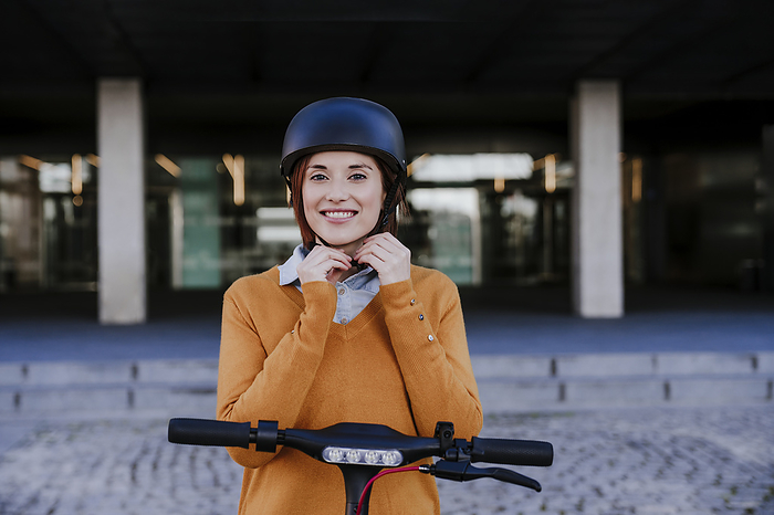 Smiling woman adjusting helmet and standing near building