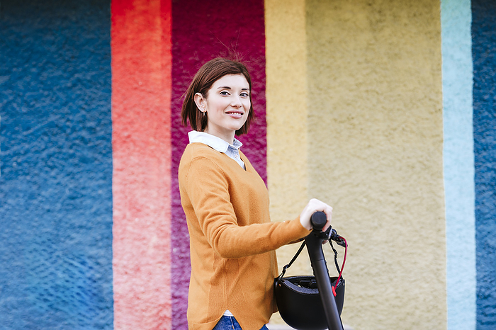 Happy woman with electric push scooter standing in front of multi colored wall