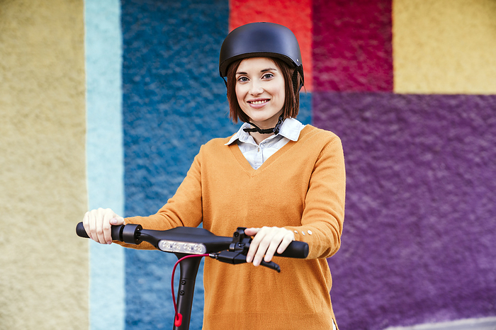 Smiling woman standing with electric push scooter in front of wall