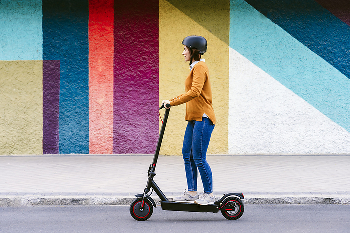 Woman wearing helmet and riding electric push scooter in front of multi colored wall