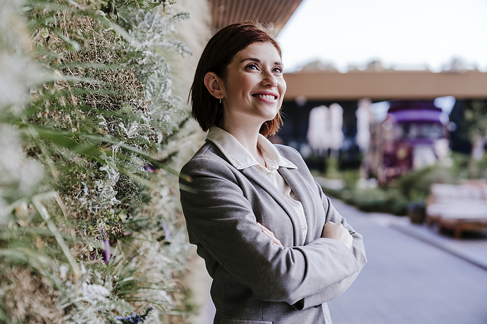 Happy businesswoman standing with arms crossed near plants