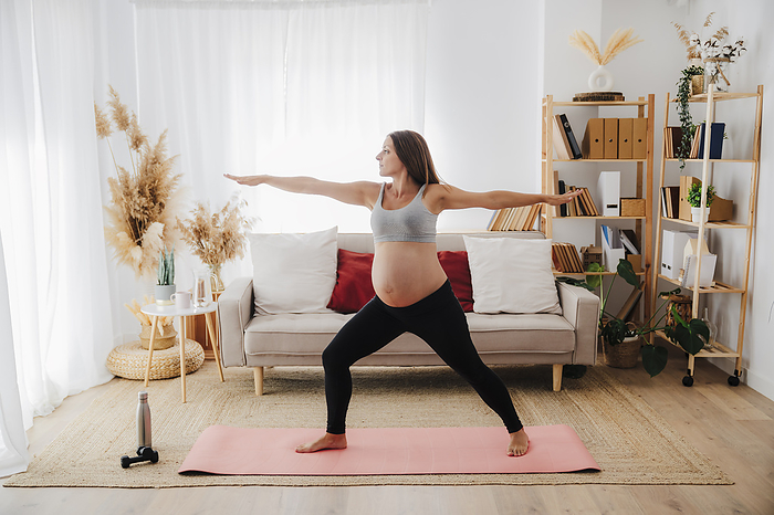 Pregnant woman practicing warrior pose yoga on mat in living room at home
