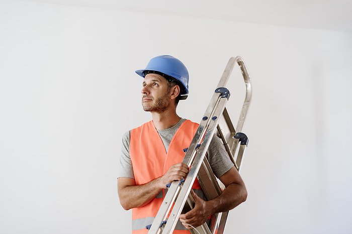 Mature technician wearing hardhat and carrying ladder at home