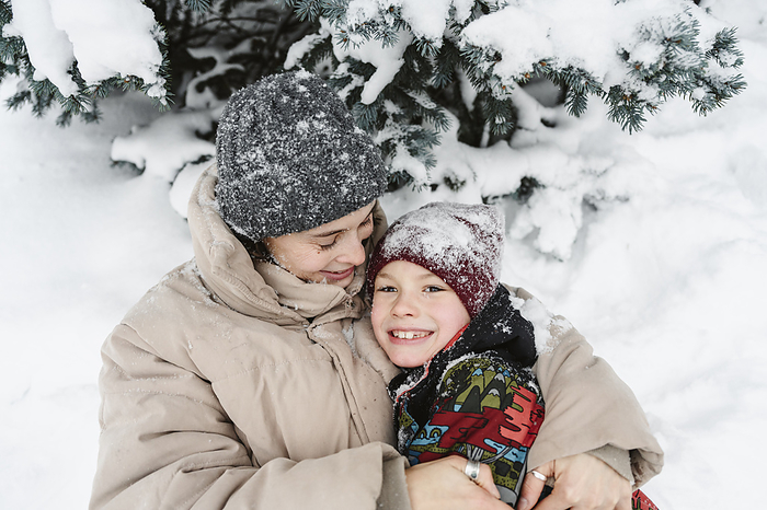 Happy woman embracing son and lying on snow near tree