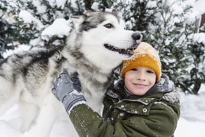 Smiling boy petting husky dog in on snow near tree