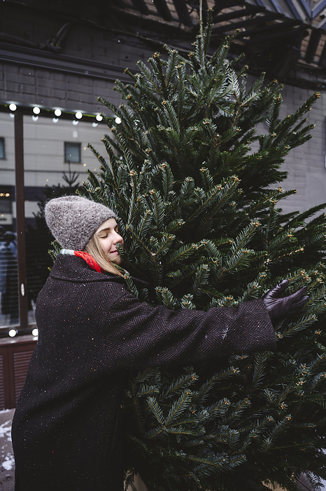 Smiling woman wearing knit hat and hugging Christmas tree