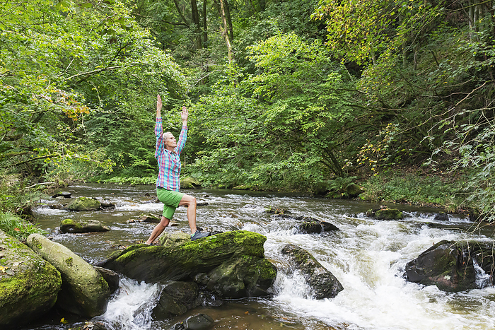 Senior woman exercising with hands raised in forest by stream