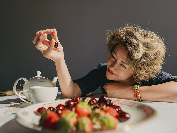 Smiling mature woman picking up cherry from plate on table at home