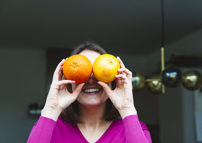 Smiling woman covering eyes with oranges at home