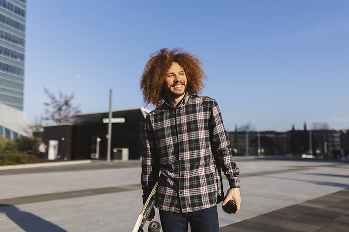 Happy man with redhead curly hair holding skateboard on sunny day
